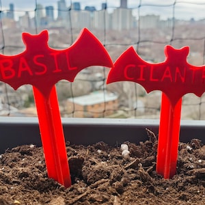 May include: Two red bat-shaped plant markers with the words "BASIL" and "CILANTRO" printed on them. The markers are stuck in the ground of a potted plant.