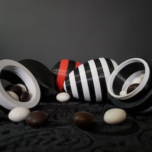 May include: A group of black and white striped, red and black, and black and white eggs with a white interior. The eggs are sitting on a black surface with chocolate sweets scattered around them.