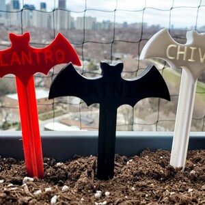 May include: Three bat-shaped plant markers, one red, one black, and one white, with the words "Cilantro", "Basil", and "Chives" printed on them.