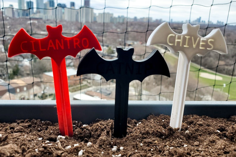 May include: Three bat-shaped plant markers, one red, one black, and one white, with the words "Cilantro", "Basil", and "Chives" printed on them.