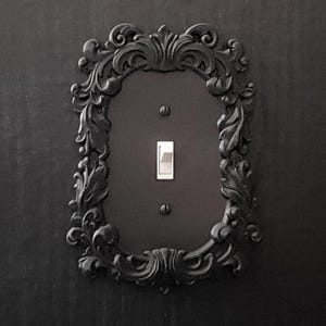 Pode incluir: Uma placa de interruptor de luz preta com uma moldura decorativa preta adornada.