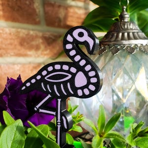 May include: A black and lavender flamingo skeleton garden stake. The flamingo has a skeleton design with a black outline and lavender interior. The garden stake is surrounded by purple flowers and green foliage.