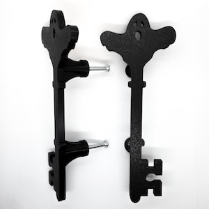 Pode incluir: Duas puxadores de armário pretos em forma de chave. Um puxador é montado com um parafuso. Os puxadores em forma de chave têm um design semelhante a um fantasma na parte superior. Os puxadores são feitos de um material preto fosco e são montados em uma superfície branca.