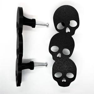 May include: Black skull-shaped cabinet knobs with a textured finish. The knobs are attached to a black vertical plate with screws. The design features three stacked skulls, creating a unique and spooky aesthetic. Ideal for adding a gothic touch to furniture.