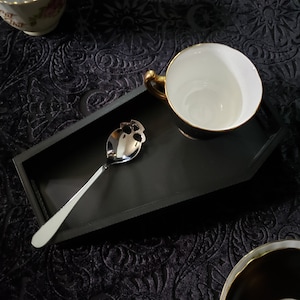 May include: A black coffin-shaped serving tray with a gold rim, a silver skull-shaped spoon, and a white teacup with a gold rim.