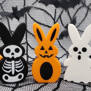 Può includere: Tre figurine di coniglio a tema Halloween. Il primo coniglio è nero con un design scheletrico bianco. Il secondo coniglio è arancione con un viso di zucca. Il terzo coniglio è bianco con un viso di fantasma.