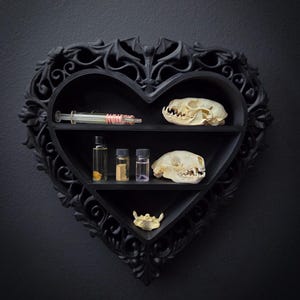 Peut inclure: Une étagère noire en forme de cœur avec des détails ornés, contenant une seringue, de petits flacons, des crânes d'animaux et un os. L'étagère est fixée à un mur gris foncé, créant une esthétique gothique ou macabre.