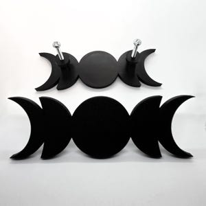 May include: Two black wooden wall hooks in a triple moon design. Each hook features a central circle flanked by crescent moons. The top hook has two silver-colored screws. The hooks are displayed against a white background.