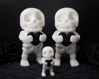 May include: Three white skeleton figurines holding black heart-shaped objects. The skeletons are different sizes, with the smallest one standing in front of the two larger ones.