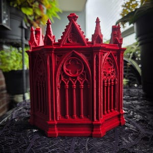 May include: A bright red, hexagonal container featuring intricate architectural details reminiscent of a Gothic cathedral. The structure includes pointed arches, vertical columns, and decorative tracery patterns along each of its six exterior sides.