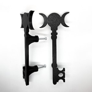May include: Two black metal key-shaped wall hooks. Each hook features a triple moon design at the top and a key-shaped bottom. The hooks are mounted with black hardware and silver screws, perfect for hanging items. The hooks are against a white background.