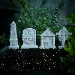 Könnte beinhalten: Vier hellgraue Gartenstecker aus Stein in Form von Miniatur-Grabsteinen. Die Stecker sind mit den Wörtern THYME, PARSLEY, OREGANO und BASIL beschriftet und zeigen jeweils ein kleines geprägtes Pflanzen- oder Totenkopf-Symbol.