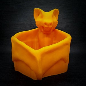 Puede incluir: Maceta naranja impresa en 3D con forma de murci&eacute;lago con una peque&ntilde;a parte superior abierta. El murci&eacute;lago tiene una boca ancha y orejas grandes.