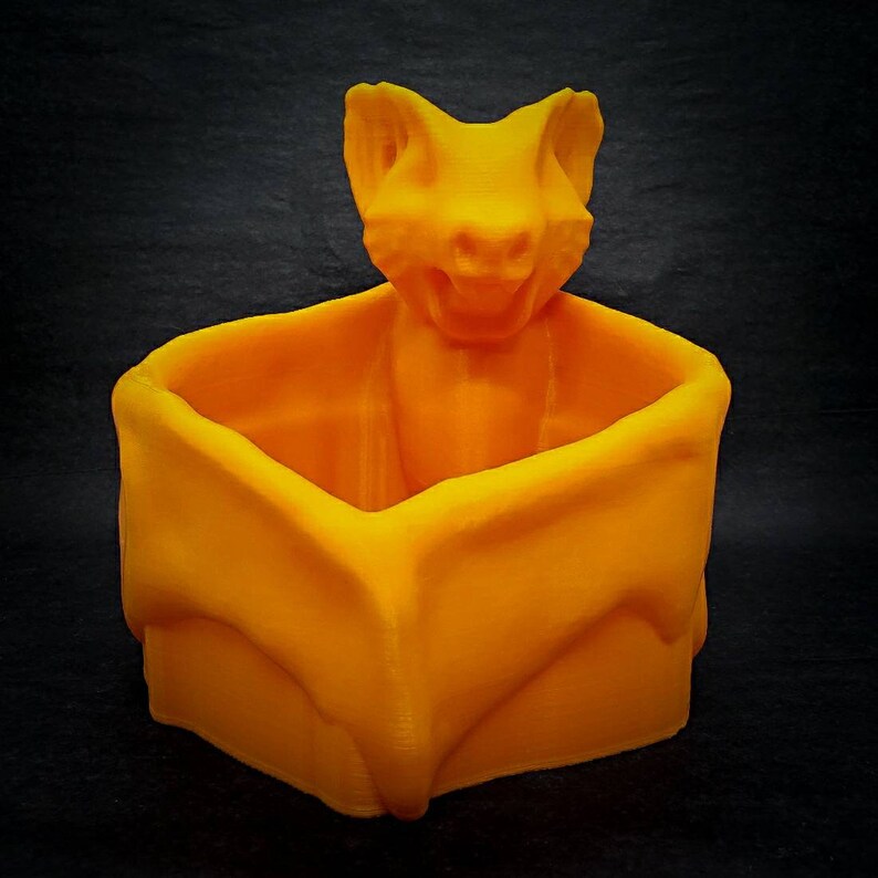 Puede incluir: Maceta naranja impresa en 3D con forma de murci&eacute;lago con una peque&ntilde;a parte superior abierta. El murci&eacute;lago tiene una boca ancha y orejas grandes.