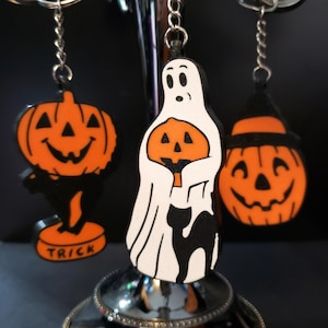 May include: Three Halloween-themed keychains: a white ghost holding an orange pumpkin, an orange pumpkin with a black cat, and an orange pumpkin with a black cat and the word "TRICK" on the base.