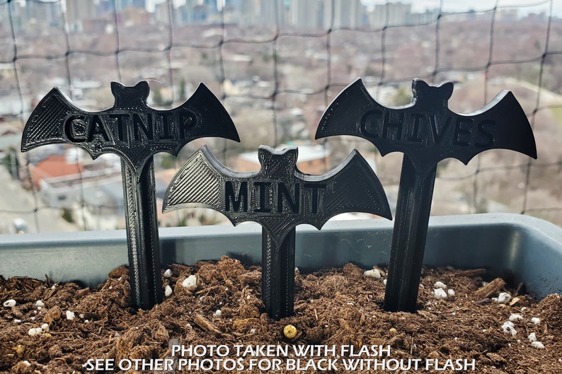 May include: Three black bat-shaped plant markers with the words "Catnip", "Mint", and "Chives" printed on them. The markers are inserted into a pot of soil.