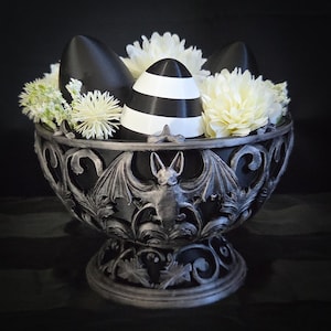 Puede incluir: Un cuenco decorativo de color plateado con un diseño de murciélago y alas, lleno de objetos en forma de huevo a rayas blancas y negras y negros sólidos, y flores blancas y verdes. El cuenco tiene un estilo gótico ornamentado.