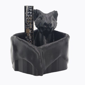 Puede incluir: Un macetero negro impreso en 3D con forma de cabeza de criatura con una regla en su interior. El macetero tiene una parte superior ancha y abierta y una parte inferior redondeada.