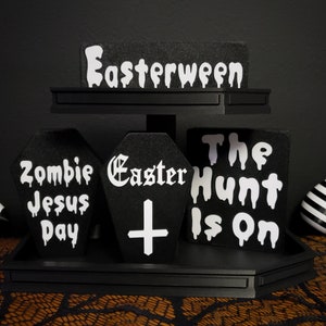 Può includere: Cartelli in legno bianco e nero con le parole "Easterween", "Zombie Jesus Day", "Easter +" e "The Hunt Is On". I cartelli sono disposti su un supporto nero a più livelli.