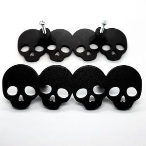 May include: Black skull-shaped decorative hardware. The hardware is made of a matte black material and features a skull design with eye and nose openings. Two sets of four skulls are shown, with screws attached to the top set.
