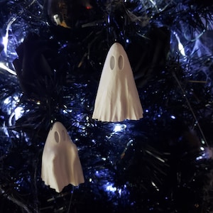 May include: Two white ghost decorations with oval eye holes hang on a dark blue Christmas tree. The tree is decorated with tinsel and lights, creating a spooky holiday theme.