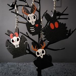 May include: Four black wooden keychains with different spooky designs. Each keychain features a skull and is hanging from a black chain. The designs include a bunny with antlers, a bat with wings, a bunny with a heart, and a creature with large eyes.