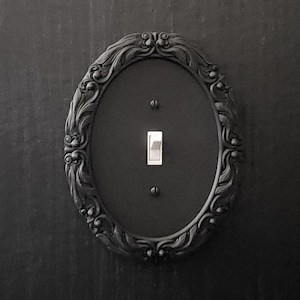 Puede incluir: Cubierta de interruptor de luz ovalada negra con detalles ornamentales de volutas. La placa del interruptor tiene un acabado mate y un interruptor basculante plateado. El diseño está enmarcado por un borde decorativo de color oscuro.