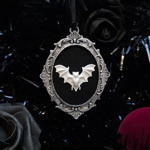 May include: A gothic-themed ornament featuring a white bat inside an ornate silver frame. The bat has outstretched wings and is centered on a black background. The ornament is suspended by a black ribbon, suitable for Halloween or gothic decor.