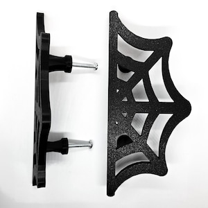 May include: Two black cabinet pulls featuring a spiderweb design. Each pull has a knob and a screw for installation. The pulls are made of a matte black material, designed for mounting on a cabinet or drawer.