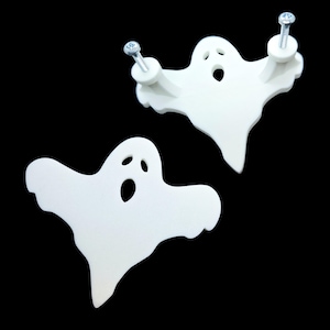 May include: Two white ghost-shaped decorative items. One ghost has two screws attached, and the other is a standalone cutout. Each ghost has three eye holes and a mouth hole, with a black background.