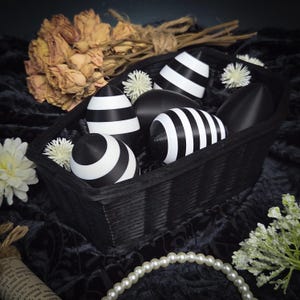 May include: A black rectangular basket containing black and white striped and solid black decorative eggs. The basket is surrounded by white flowers, dried flowers, and a string of pearls. The background is a dark, textured fabric.