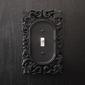 Puede incluir: Una placa de interruptor de luz negra con un marco ornamentado y decorativo. El marco est&aacute; hecho de un material negro texturizado y tiene un dise&ntilde;o detallado en forma de rollo.