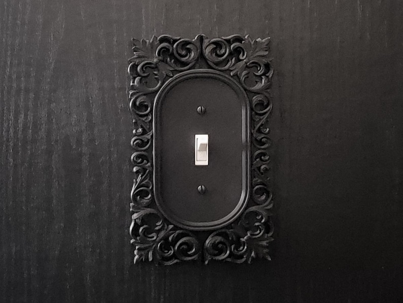 Puede incluir: Una placa de interruptor de luz negra con un marco ornamentado y decorativo. El marco est&aacute; hecho de un material negro texturizado y tiene un dise&ntilde;o detallado en forma de rollo.