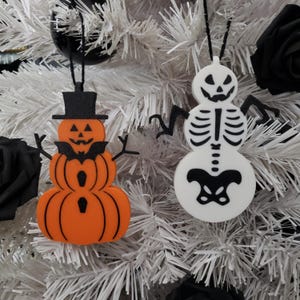 以下が含まれることがあります： 雪だるまの形をしたハロウィーンの飾り2つ。1つの雪だるまはオレンジ色で、かぼちゃの顔と黒いシルクハットをかぶっています。もう1つの雪だるまは白で、骸骨の顔と骨を持っています。