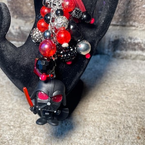 May include: A keychain featuring a small Darth Vader figure holding a red lightsaber, with red, black, and silver beads. The beads are various sizes and textures, including faceted, glittery, and smooth. A red tassel is also included.