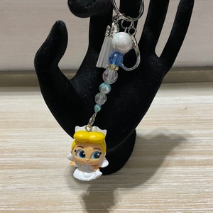 May include: A handmade keychain featuring a small Cinderella figurine in a white dress. The keychain has a silver-tone metal ring, a white tassel, and a strand of blue and clear beads. The figurine has blonde hair and blue eyes.