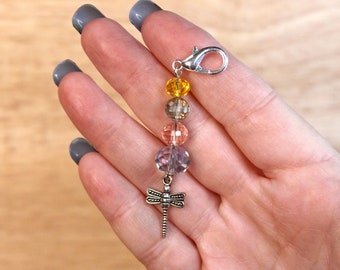 May include: A silver charm with a dragonfly pendant and a series of clear glass beads in shades of yellow, orange, and pink. The charm is attached to a silver lobster clasp.