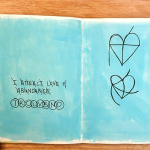 May include: An open notebook with a blue background. The left page has the text "I attract love & abundance" written in black ink. The right page has two heart symbols drawn in black ink.