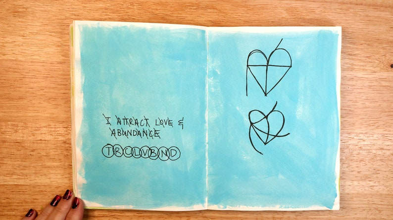 May include: An open notebook with a blue background. The left page has the text "I attract love & abundance" written in black ink. The right page has two heart symbols drawn in black ink.
