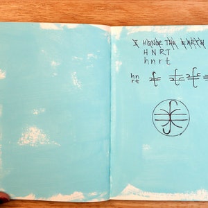 May include: An open notebook with a light blue cover. The right page has handwritten text that says "I honor the Earth" and a symbol with a circle and lines.