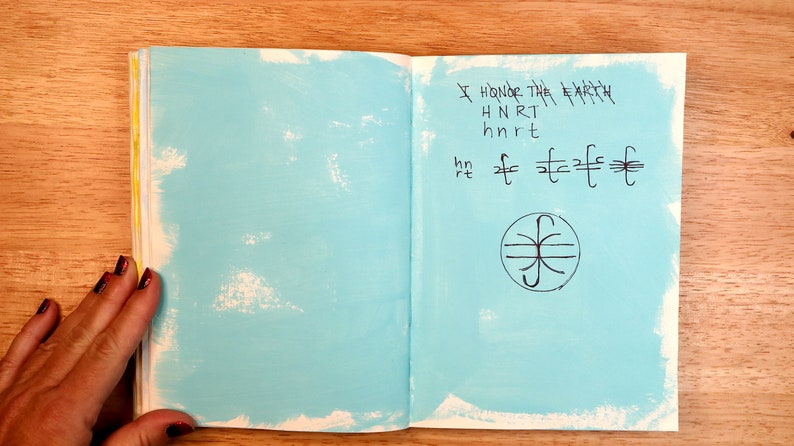 May include: An open notebook with a light blue cover. The right page has handwritten text that says "I honor the Earth" and a symbol with a circle and lines.