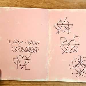 May include: An open notebook with pink pages and black ink drawings of hearts. The text "I draw love in drawn" is written on the left page.