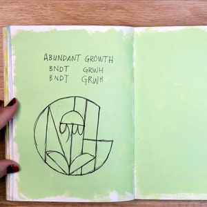 May include: An open notebook with a green page on the right and a green page on the left with black ink. The left page has the words "ABUNDANT GROWTH" written at the top and the words "BNDT GRWH" written twice below. A black and white drawing of a stylized flower is in the center of the page.