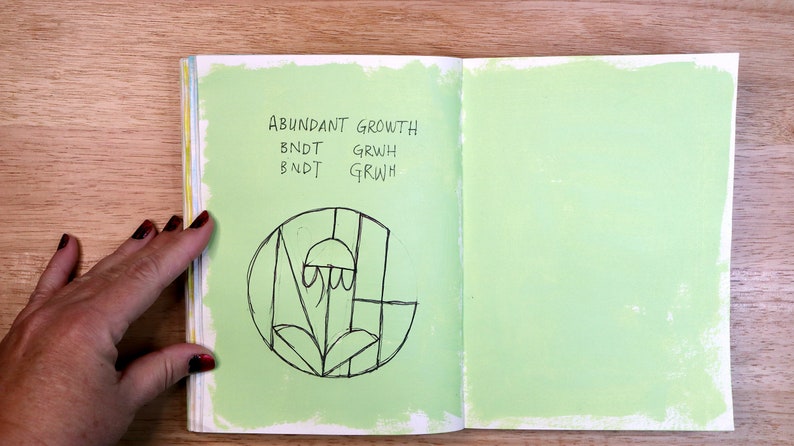 May include: An open notebook with a green page on the right and a green page on the left with black ink. The left page has the words "ABUNDANT GROWTH" written at the top and the words "BNDT GRWH" written twice below. A black and white drawing of a stylized flower is in the center of the page.