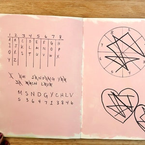 May include: A pink notebook with a gridded alphabet chart, a message written in black ink, and a circle with lines drawn through it. The notebook also has two hearts with lines drawn through them.