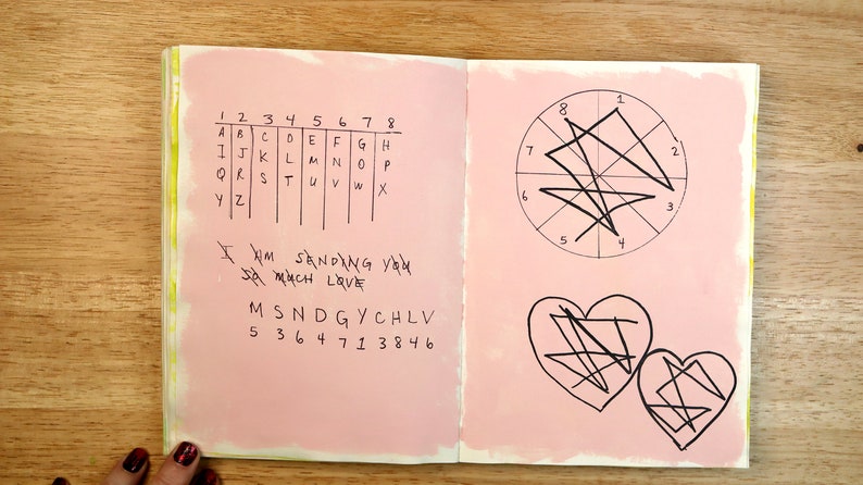 May include: A pink notebook with a gridded alphabet chart, a message written in black ink, and a circle with lines drawn through it. The notebook also has two hearts with lines drawn through them.