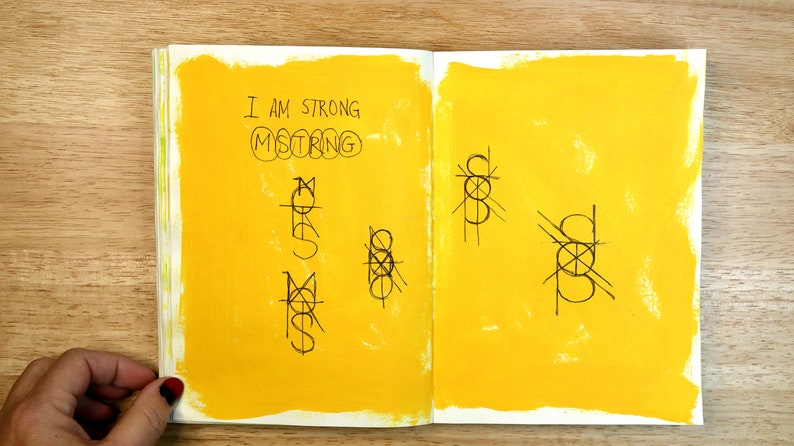 May include: A yellow notebook with the words "I am strong" and "mistring" written on the left page. The right page has a series of abstract drawings.