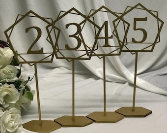 May include: Four gold geometric table number holders with the numbers 2, 3, 4, and 5 displayed on them. The holders are on a white surface.