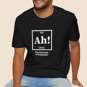 May include: Black t-shirt featuring a white graphic with the word "Ah!" in a square, and the text "The Element of Surprise!" below. The shirt is paired with blue jeans.