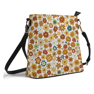 May include: A crossbody bag featuring a white base with a dense, repeating pattern of small flowers in shades of yellow, orange, brown, and teal. The bag includes a black adjustable shoulder strap attached with silver-toned metal hardware.